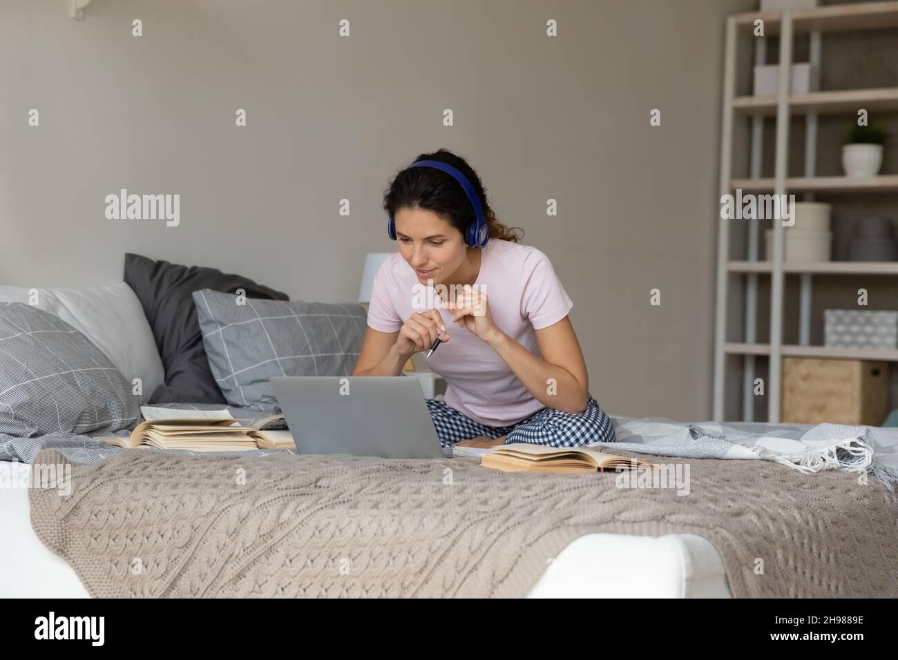 Nachdenkliche Frau in kabellosen Kopfhörern, die online studiert und auf dem Bett sitzt Stockfoto