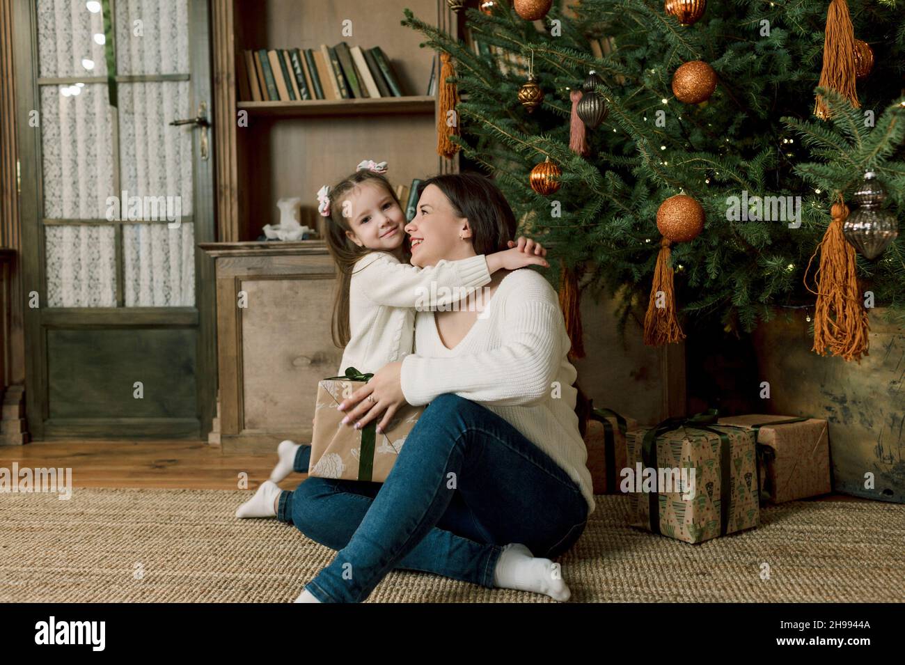 Eine glückliche Familie, die sich über Weihnachten freut. Eine lächelnde Tochter umarmt ihre Mutter im Wohnzimmer mit einem geschmückten Weihnachtsbaum im Bac Stockfoto