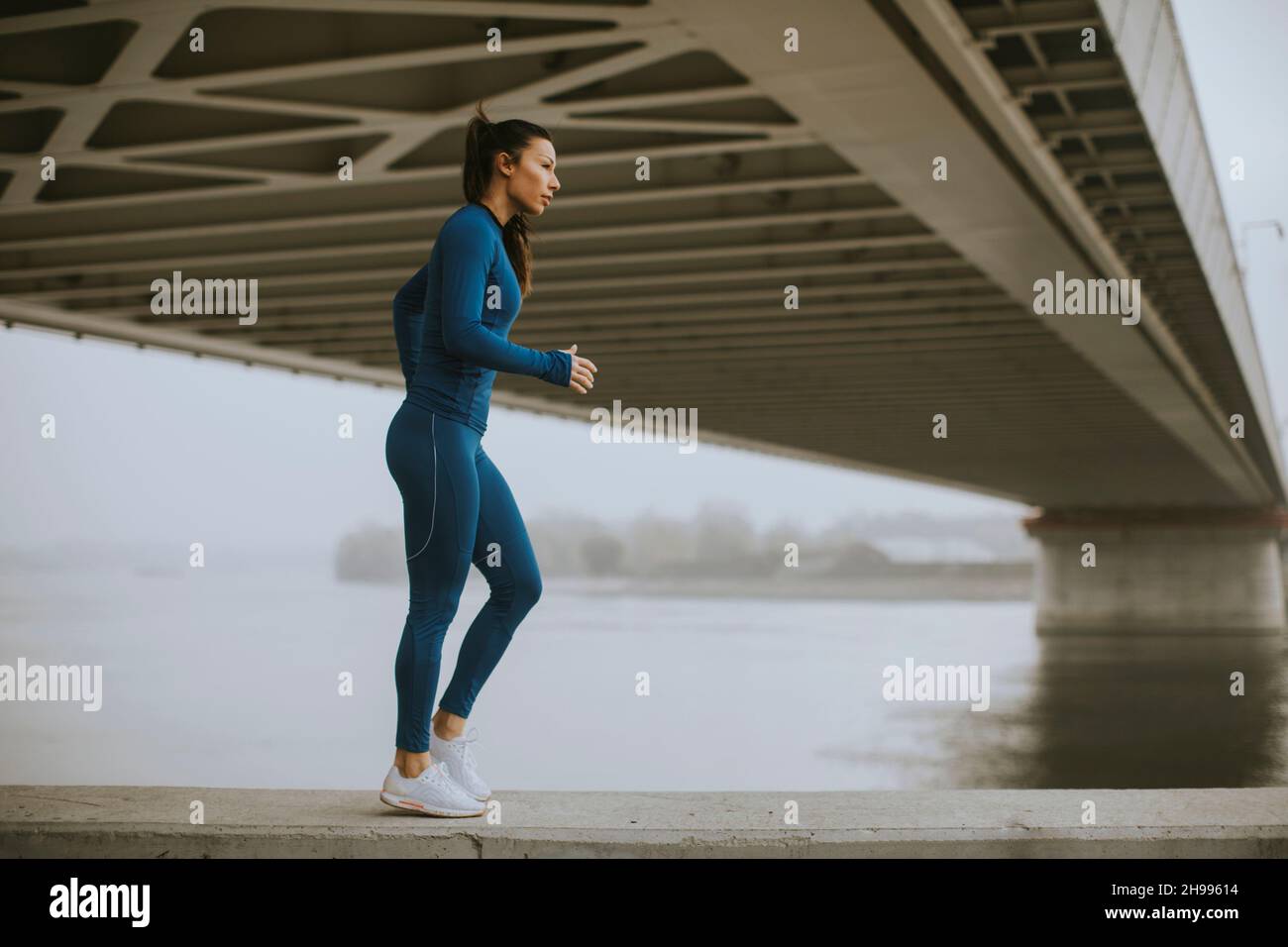 Hübsche junge Frau im blauen Trainingsanzug, die sich vor dem Training am Fluss am Herbstmorgen ausdehnt Stockfoto