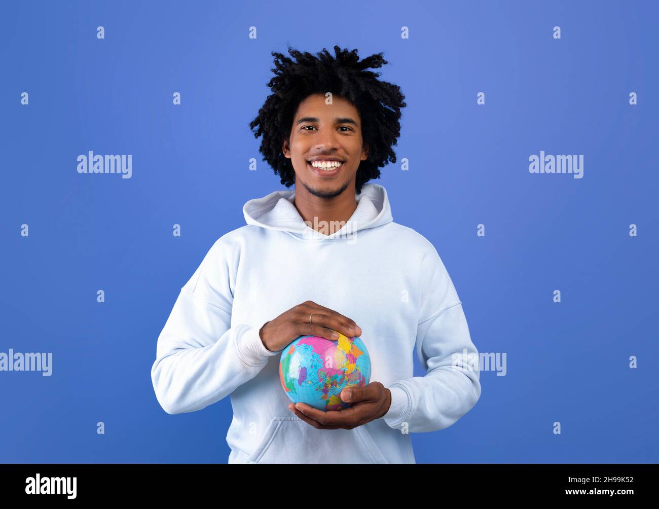 Glücklicher schwarzer Teenager, der den Globus mit seinen Händen auf blauem Studiohintergrund beschützt. Ökologie, Tourismus, Gleichheit der Menschheit Stockfoto