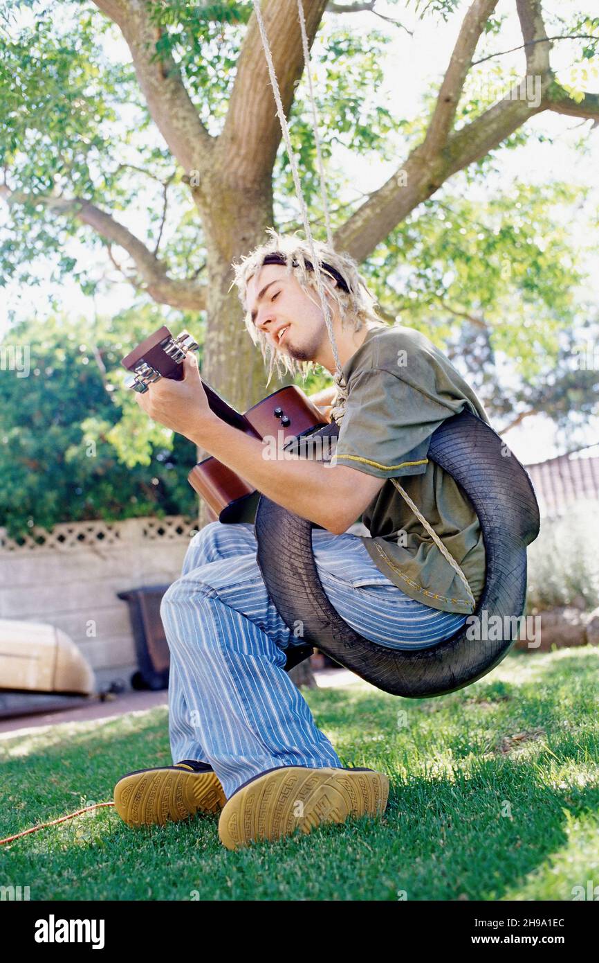 Mann, der Gitarre spielt, sitzt auf einer Reifenschaukel in einem Garten und singt. Stockfoto