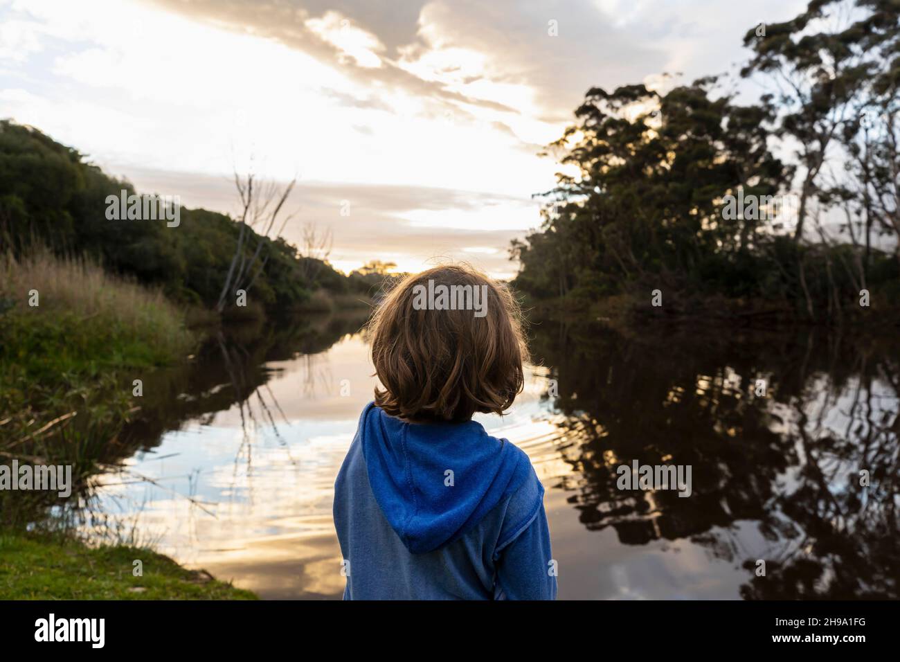 Ein kleiner Junge, der in der Dämmerung an einem Fluss steht, reflektiert den Himmel im flachen, ruhigen Wasser Stockfoto