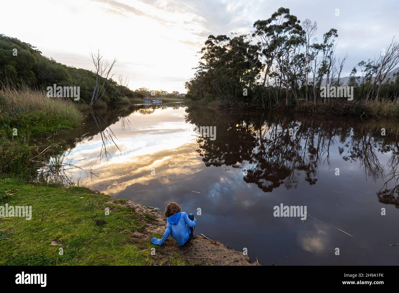 Ein kleiner Junge, der in der Dämmerung an einem Fluss steht, reflektiert den Himmel im flachen, ruhigen Wasser Stockfoto