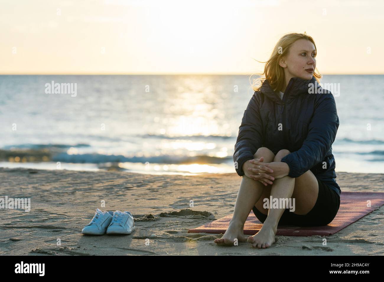 Frau, die während des Sonnenuntergangs auf einer Yogamatte mit entspanntem Ausdruck am Strand sitzt Stockfoto