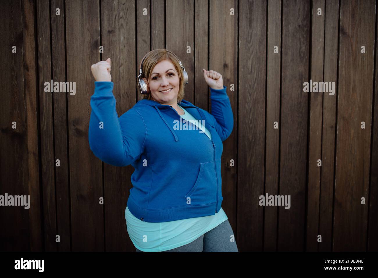 Glückliche übergewichtige Frau in Sportkleidung mit Kopfhörern und Blick auf die Kamera gegen die Holzwand Stockfoto