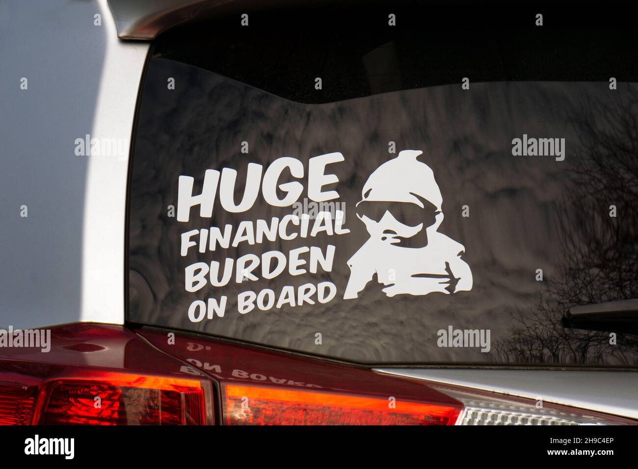 Ein Aufkleber auf der Rückseite eines Autos, der eine enorme finanzielle Belastung an Bord sagt, anstatt Baby an Bord. Auf einem Parkplatz in Queens, New York City. Stockfoto