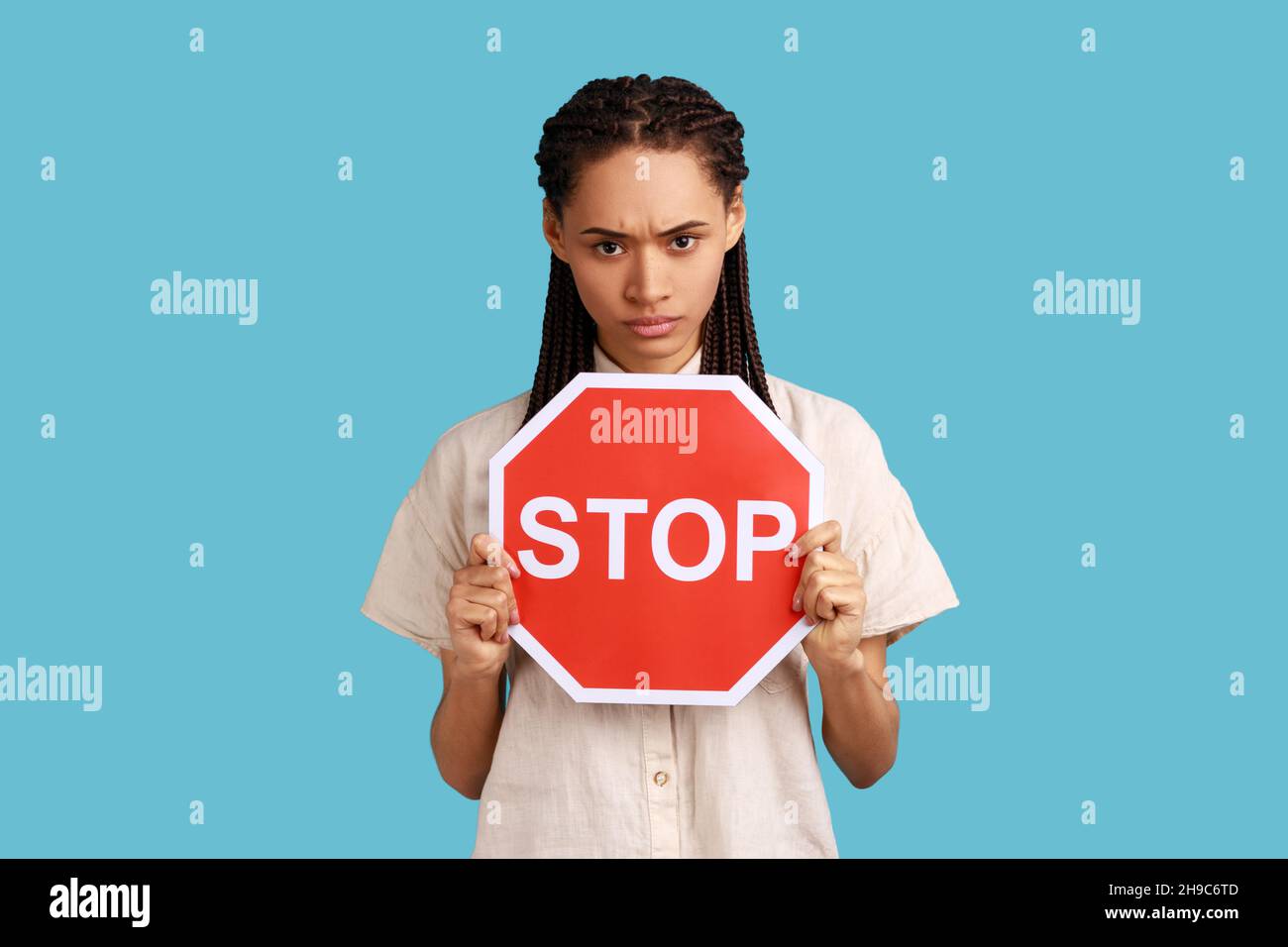 Porträt einer wütenden Frau mit Dreadlocks, die das rote Stoppschild halten, die Kamera mit negativem aggressiven Ausdruck betrachten, Verbotszeichen zeigen und ein weißes Hemd tragen. Innenaufnahme des Studios isoliert auf blauem Hintergrund Stockfoto