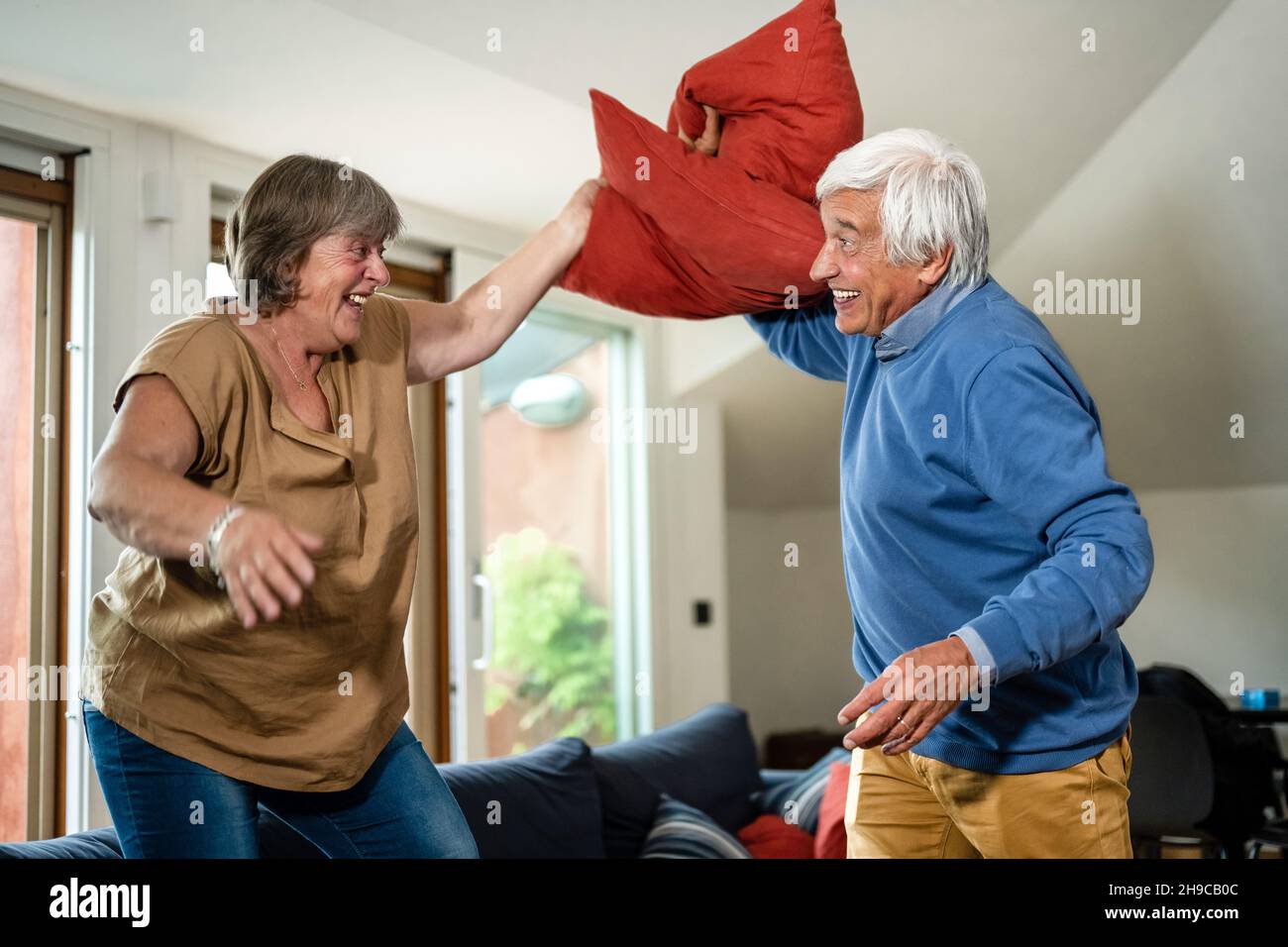 Spielerisches und sorgloses Seniorenpaar, das Kissen macht, kämpft mitten im Wohnzimmer, Freizeit-, Lifestyle- und Glückskonzept im Silberzeitalter Stockfoto
