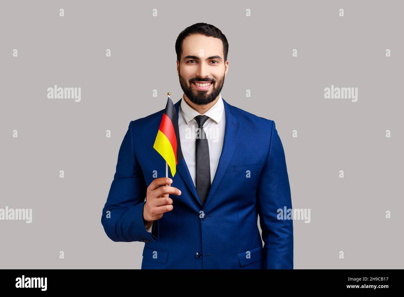 Hochgejubelter Geschäftsmann mit der deutschen Flagge, der den Tag der Bundesrepublik Deutschland - den 3th. Oktober - feiert, positive Emotionen ausdrückt und einen offiziellen Anzug trägt. Innenaufnahme des Studios isoliert auf grauem Hintergrund. Stockfoto