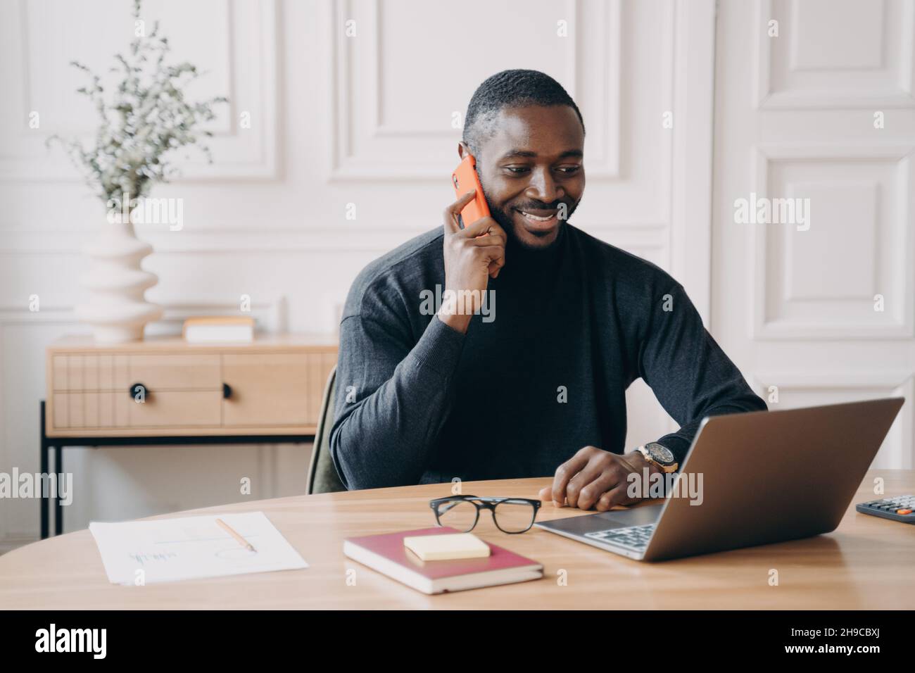 Erfolgreicher afro amerikanischer Geschäftsmann, der angenehme Telefongespräche während der Arbeit am Laptop hat Stockfoto