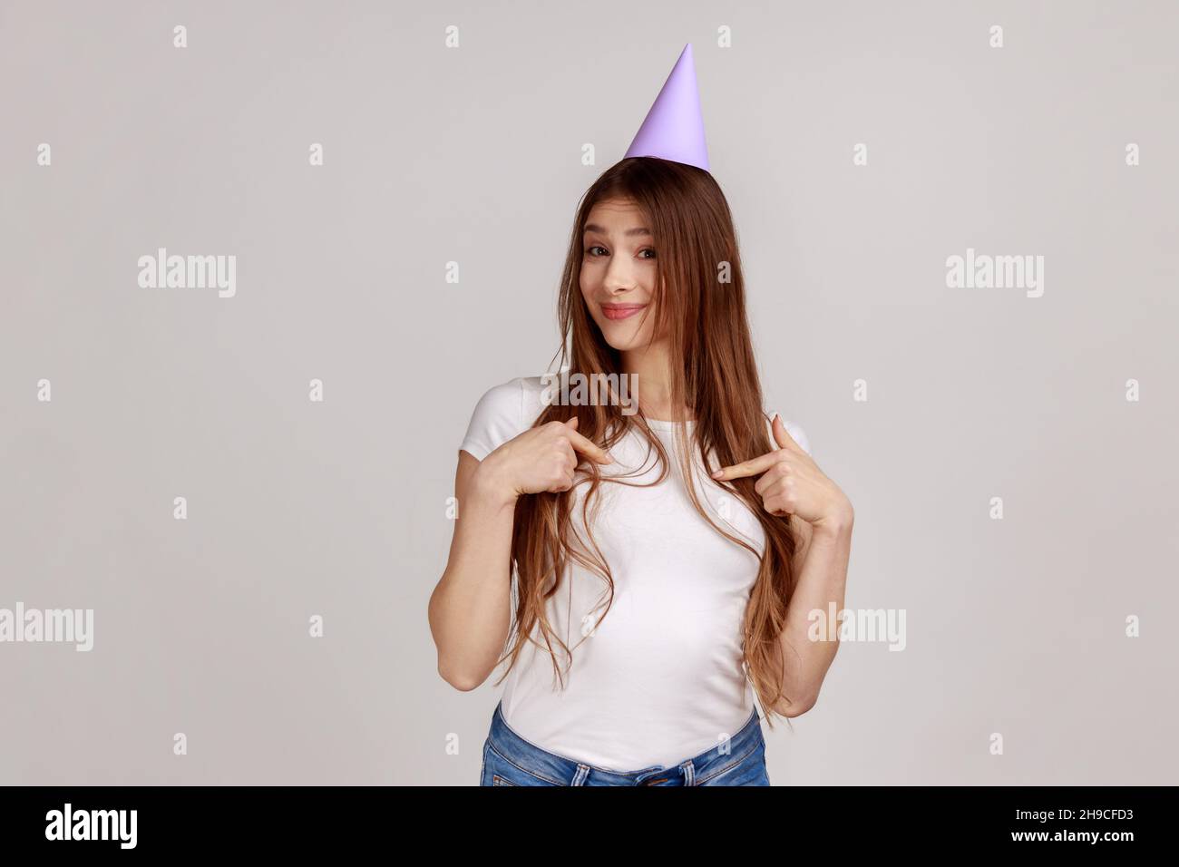 Porträt einer charmanten, fröhlichen jungen Frau in einem Partykegel auf ihrem Kopf, auf sich selbst zeigend, Geburtstag feiert und weißes T-Shirt trägt. Innenaufnahme des Studios isoliert auf grauem Hintergrund. Stockfoto