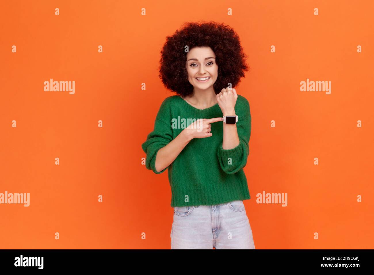 Gut aussehende positive Frau mit Afro-Frisur, die einen grünen legeren Pullover trägt, der auf die Armbanduhr zeigt und lächelt, die Uhr zeigt, Happy Hours. Innenaufnahme des Studios isoliert auf orangefarbenem Hintergrund. Stockfoto