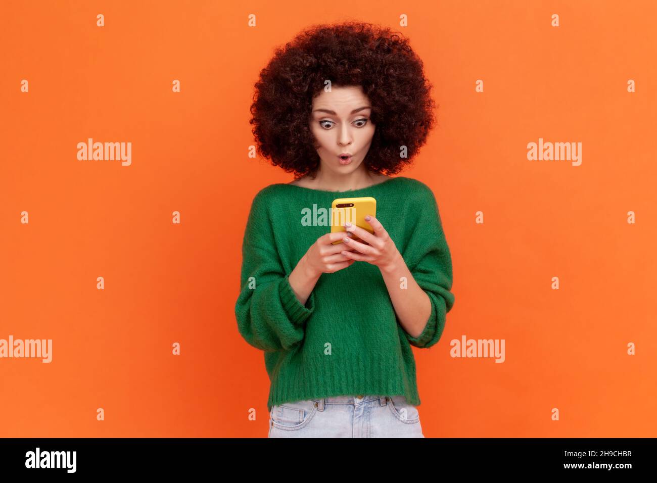 Schockierte Frau mit Afro-Frisur, die einen grünen Pullover im lässigen Stil trägt, der mit offenem Mund steht, ein Handy benutzt und erstaunliche Nachrichten in E-Mails liest. Innenaufnahme des Studios isoliert auf orangefarbenem Hintergrund. Stockfoto