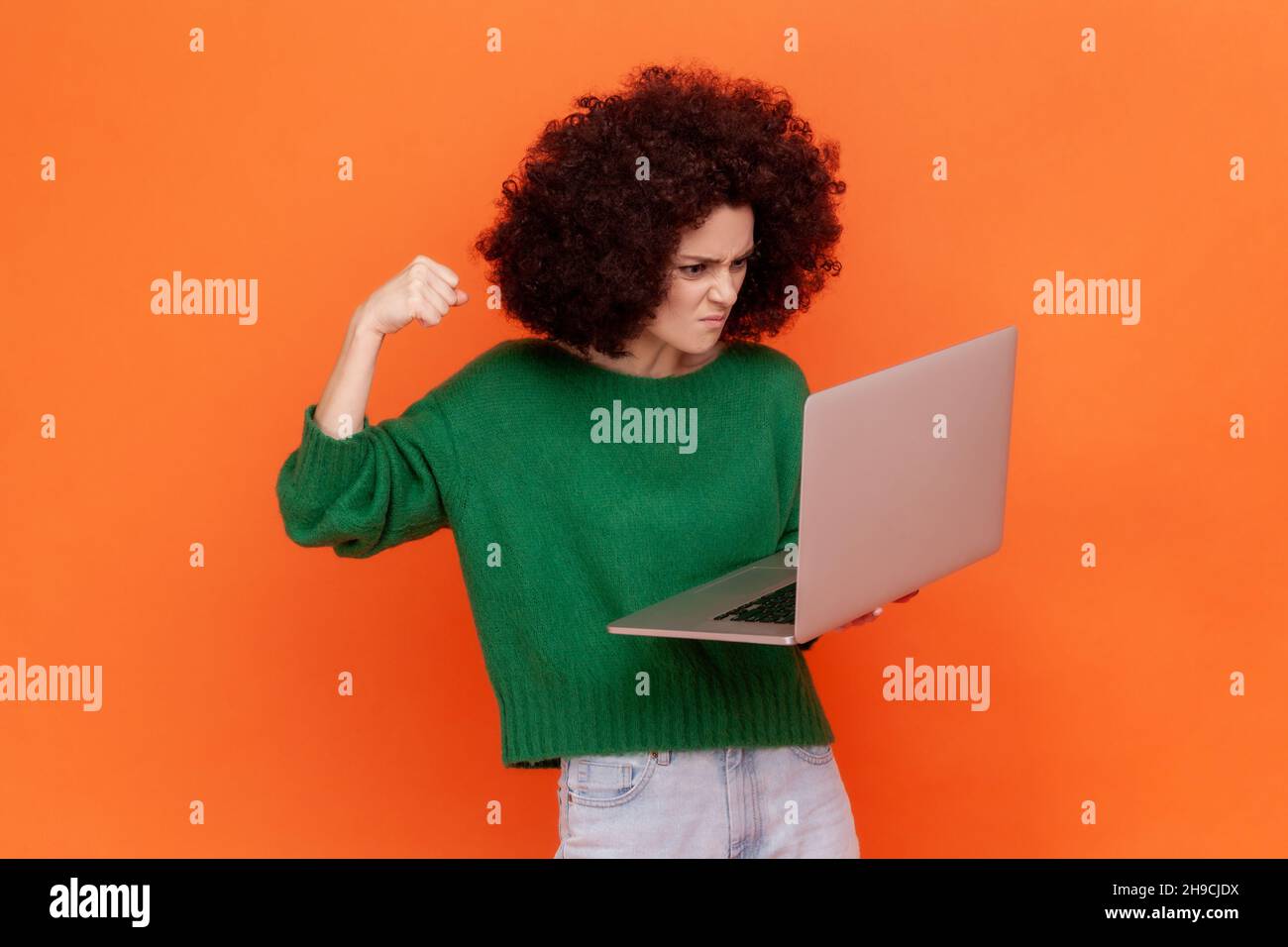 Wütende Frau mit Afro-Frisur trägt einen grünen Pullover im lässigen Stil, der am Laptop arbeitet, hat Fehler und will ihre Faust auf den Bildschirm schlagen. Innenaufnahme des Studios isoliert auf orangefarbenem Hintergrund. Stockfoto