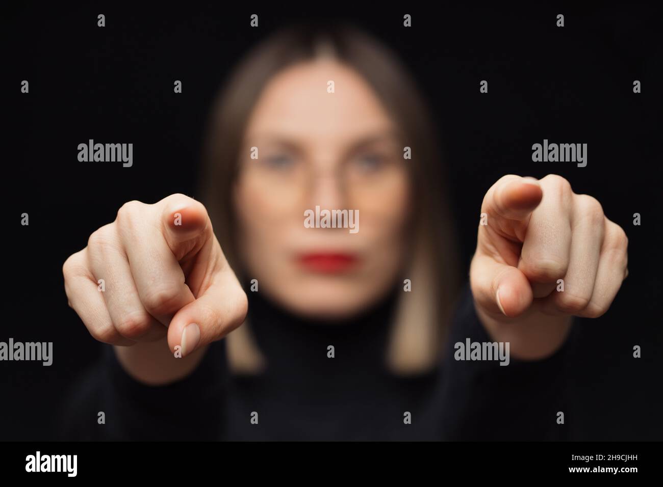 Nahaufnahme eines Frauenporträts, das die Finger auf die Kamera zeigt, mit Brille, roten Lippen, auf schwarzem Hintergrund und fokussiertem Gesicht an den Fingern. Du bist der Gewinner, ich werde dich finden. Wir suchen Sie Stockfoto