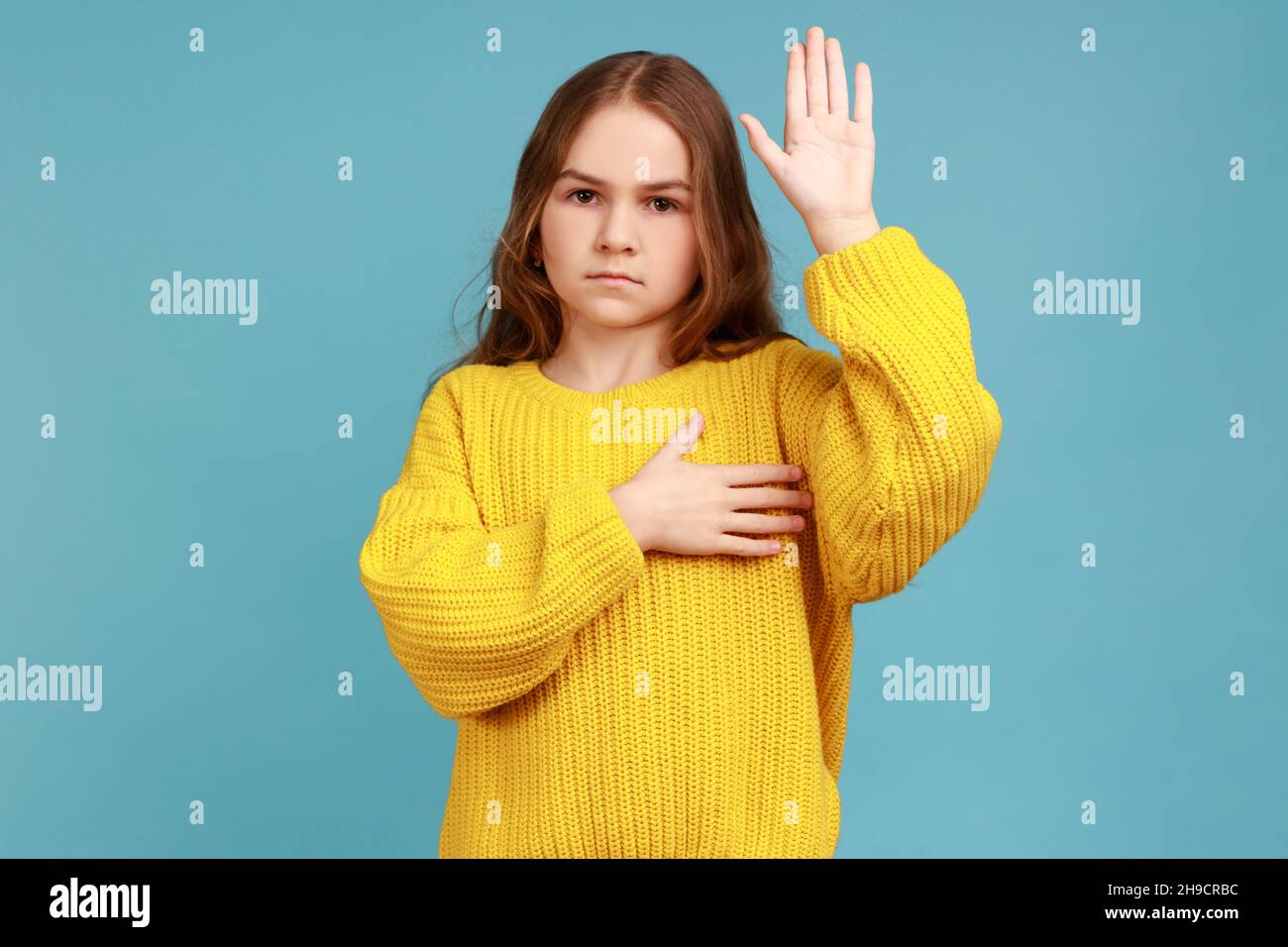 Porträt eines ernsthaften kleinen Mädchens, das zum Eid die Handfläche hebt, und eines Kindes, das sich beschworen hat, nur die Wahrheit zu sagen, und einen gelben Pullover im lässigen Stil trägt. Innenaufnahme des Studios isoliert auf blauem Hintergrund. Stockfoto