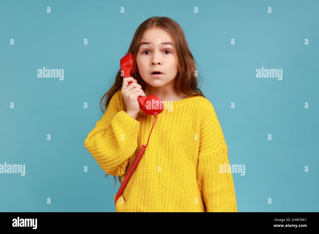 Portrait of Little girl hält Telefon Hörer überrascht von der Unterhaltung, schockiert durch unglaubliche Nachrichten, tragen gelben lässigen Stil Pullover. Innenaufnahme des Studios isoliert auf blauem Hintergrund. Stockfoto