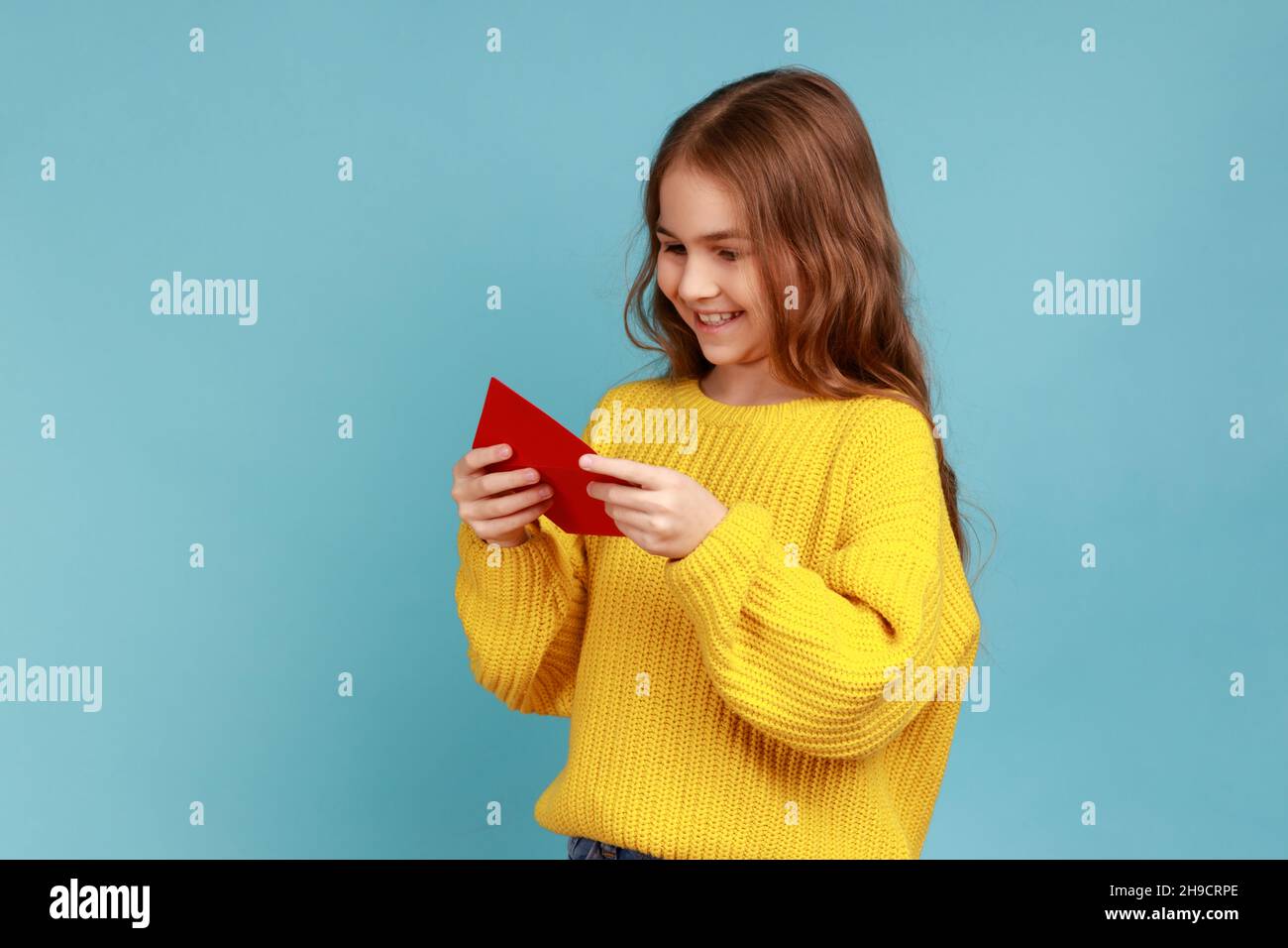 Portrait of Little girl hält roten Umschlag in den Händen, liest Brief von Klassenkamerad mit charmanten Lächeln, trägt gelben lässigen Stil Pullover. Innenaufnahme des Studios isoliert auf blauem Hintergrund. Stockfoto