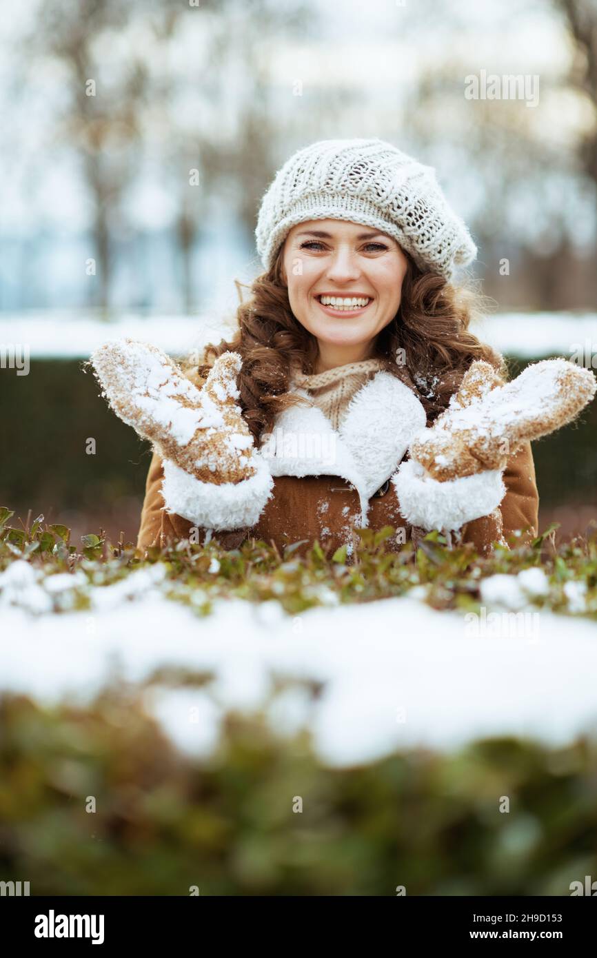 Fröhliche elegante Frau draußen im Stadtpark im Winter in einem gestrickten Hut und Schaffell Mantel mit verschneiten Fäustlingen. Stockfoto
