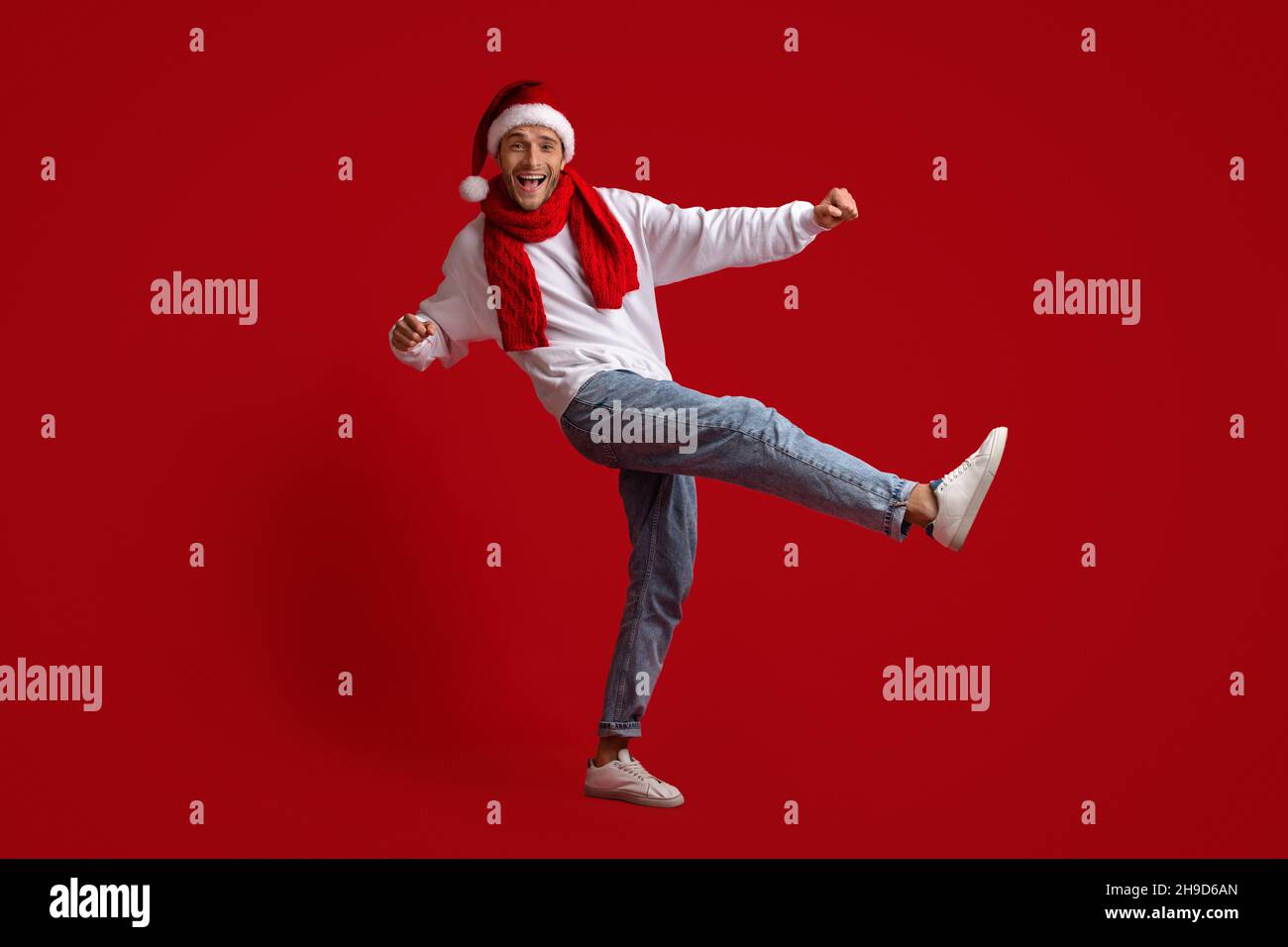 Fröhlicher Junger Mann Mit Weihnachtsmütze Und Rotem Schal, Der Im Studio Tanzt Stockfoto