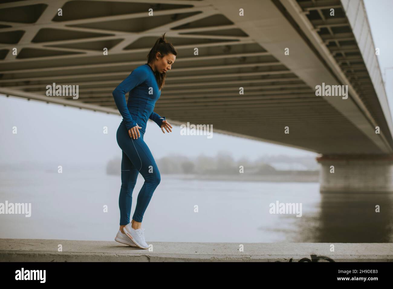 Hübsche junge Frau im blauen Trainingsanzug, die sich vor dem Training am Fluss am Herbstmorgen ausdehnt Stockfoto