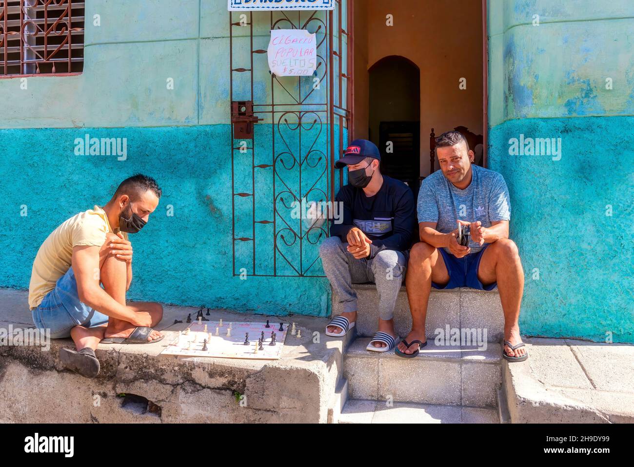 Drei kubanische Männer, die auf dem Bürgersteig Schach spielen, an einem Schild mit der Aufschrift 'Wir verkaufen Zigaretten'.Dez. 6, 2021 Stockfoto