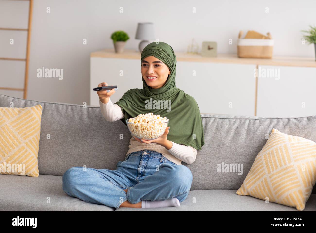 Junge arabische Frau im Hijab, die mit Fernbedienung und Popcorn fernsieht und zu Hause mit gekreuzten Beinen auf der Couch sitzt Stockfoto