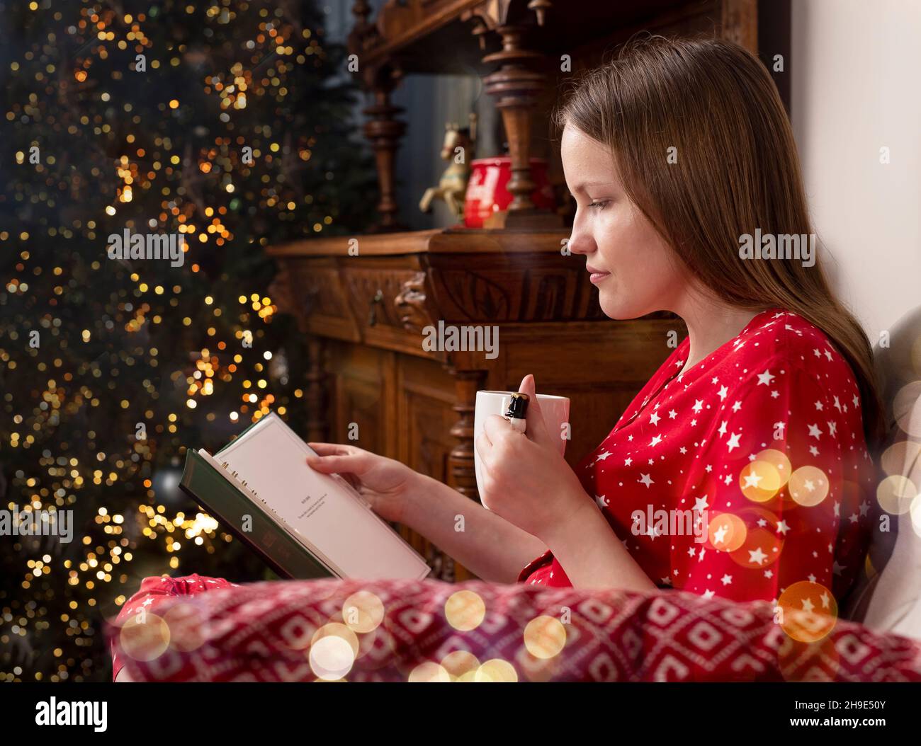Frau liest Buch, sitzt auf einem Stuhl mit Teetasse. Person entspannt mit Weihnachtsbaum auf verschwommenem Hintergrund in den Winterferien. Person, die abends im gemütlichen Zuhause liest. Stockfoto