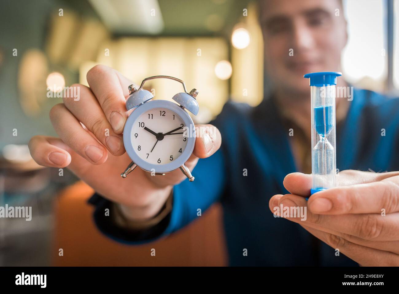 Die Hand eines männlichen Geschäftsmannes hält eine Weckzeit und eine Sanduhr, Nahaufnahme. Stockfoto