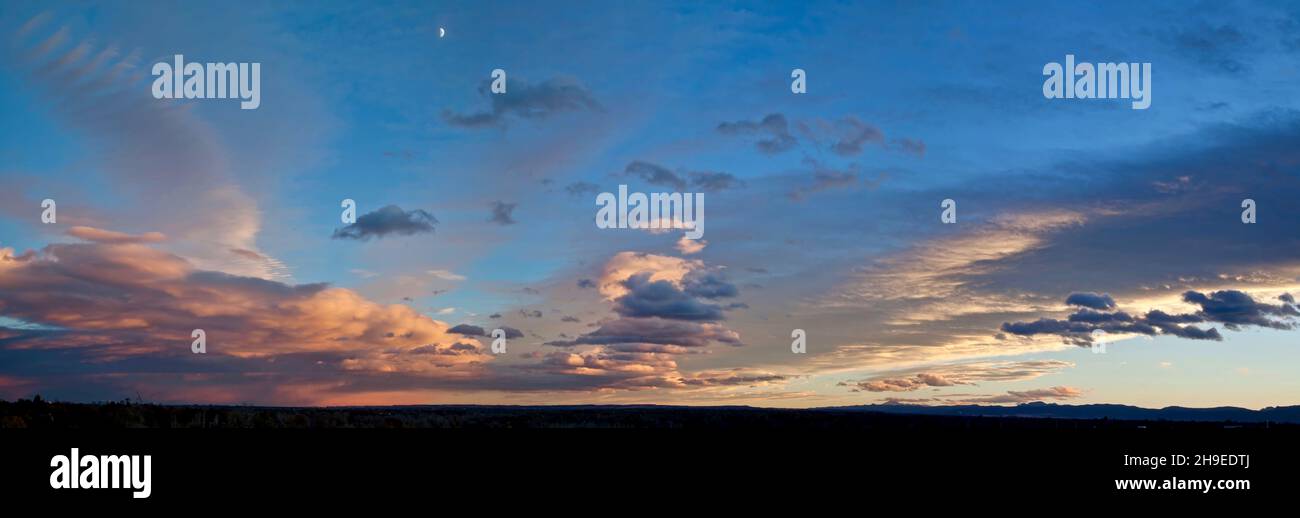 Die untergehende Sonne hebt Wolken direkt über dem Horizont und unter dem frühen Halbmond am Abend hervor. Stockfoto