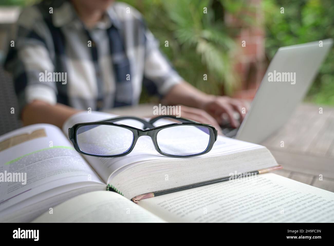 Bücher auf dem Tisch mit einer Frau und einem Computer-Laptop im Hintergrund. Selektiver Fokus auf Brillen. Stockfoto