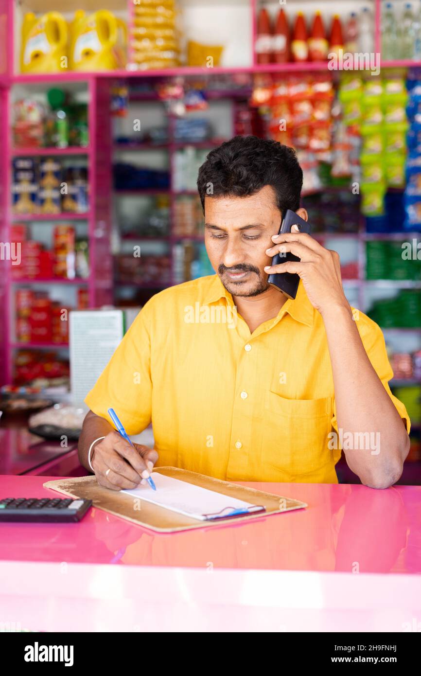 Kirana Marchant oder Eigentümer nimmt Bestellungen durch Notieren von Kunden über Handy-Anruf im Lebensmittelgeschäft - Konzept der kleinen Unternehmen und Lieferung nach Hause Stockfoto