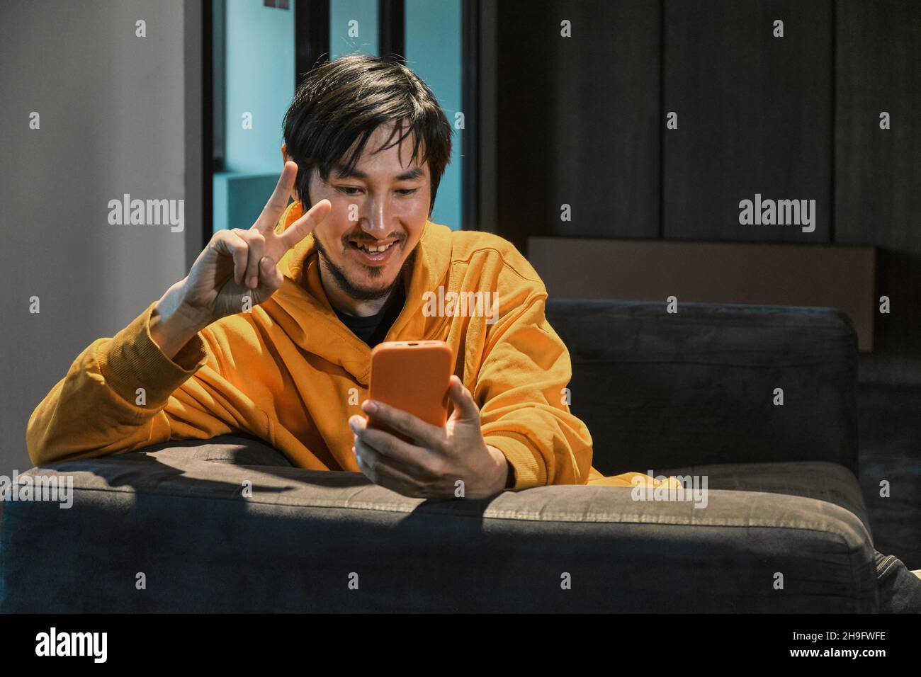 Ein Asiat sitzt in einem kleinen Büro auf dem Sofa und kommuniziert per Mobiltelefon. Das Konzept von Kleinunternehmen und Online-Kommunikation. Stockfoto