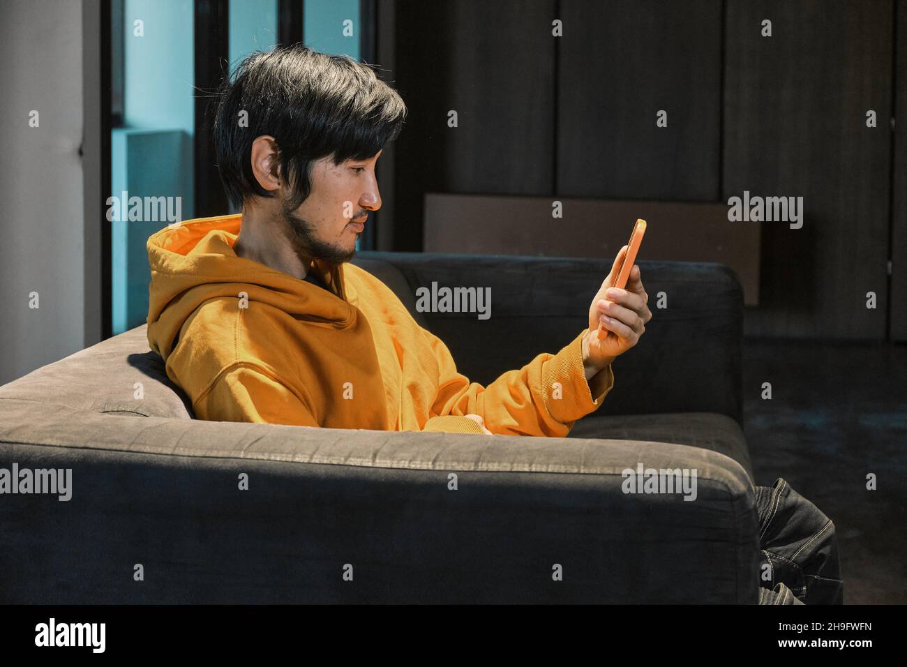 Ein Asiat sitzt in einem kleinen Büro auf dem Sofa und kommuniziert per Mobiltelefon. Das Konzept von Kleinunternehmen und Online-Kommunikation. Stockfoto