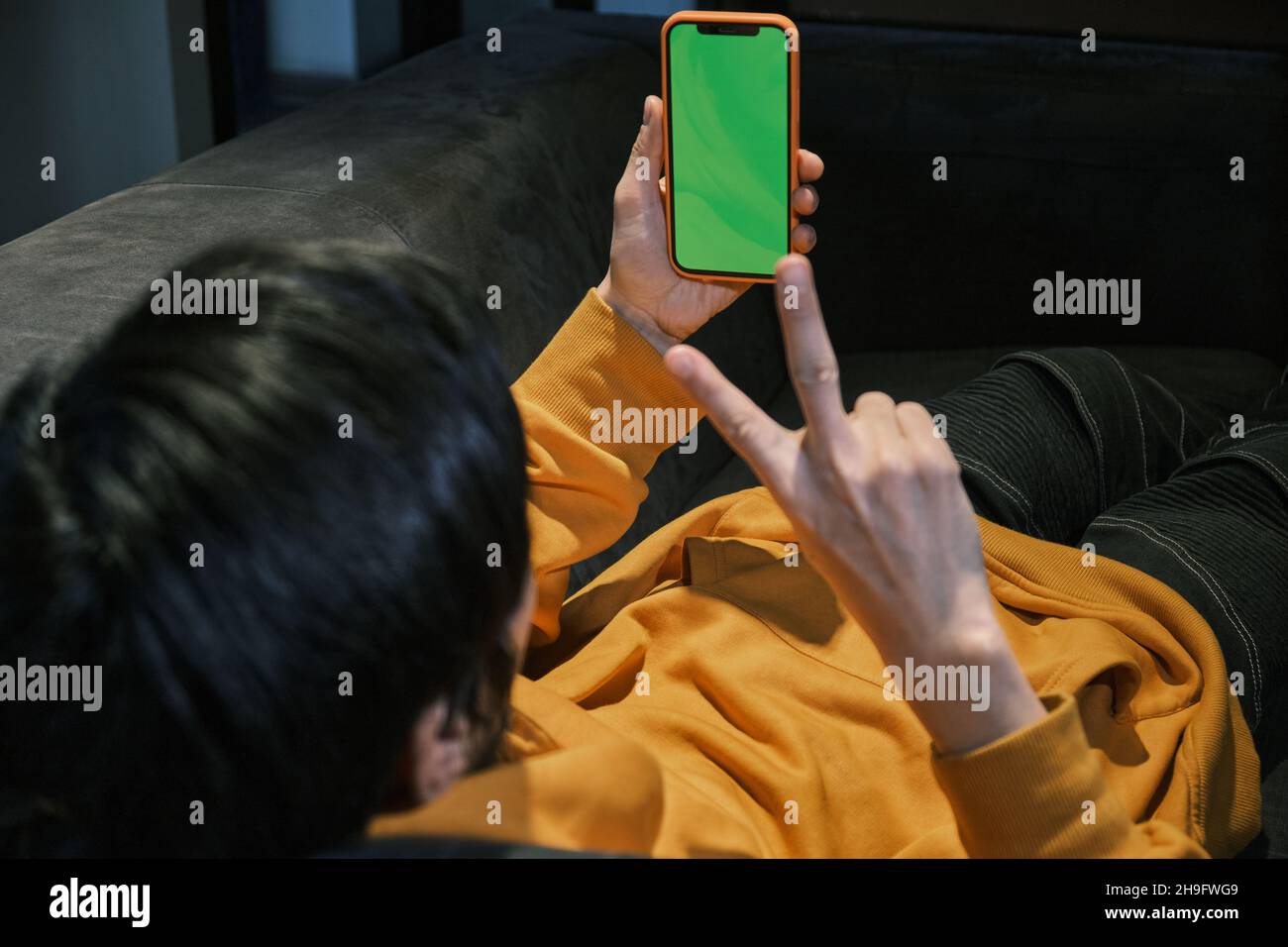 Ein asiatischer Mann legt sich in einem kleinen Büro auf das Sofa und kommuniziert via Videoverbindung über ein Mobiltelefon mit grünem Bildschirm. Das Konzept des kleinen Unternehmens und Stockfoto