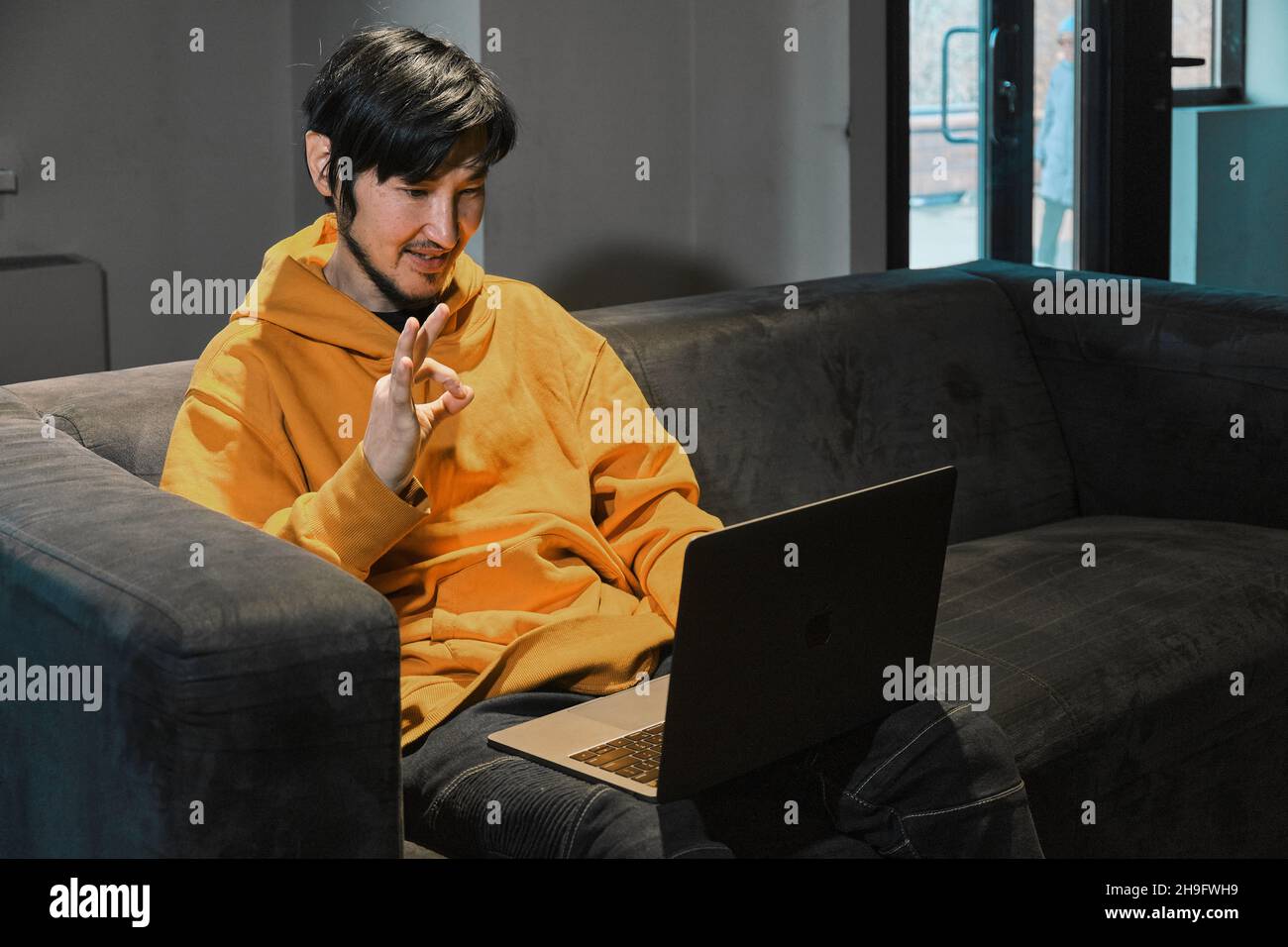 Ein Asiat sitzt in einem kleinen Büro auf dem Sofa und kommuniziert über einen Laptop über eine Videoverbindung. Das Konzept von Kleinunternehmen und Online-Kommunikation. Stockfoto