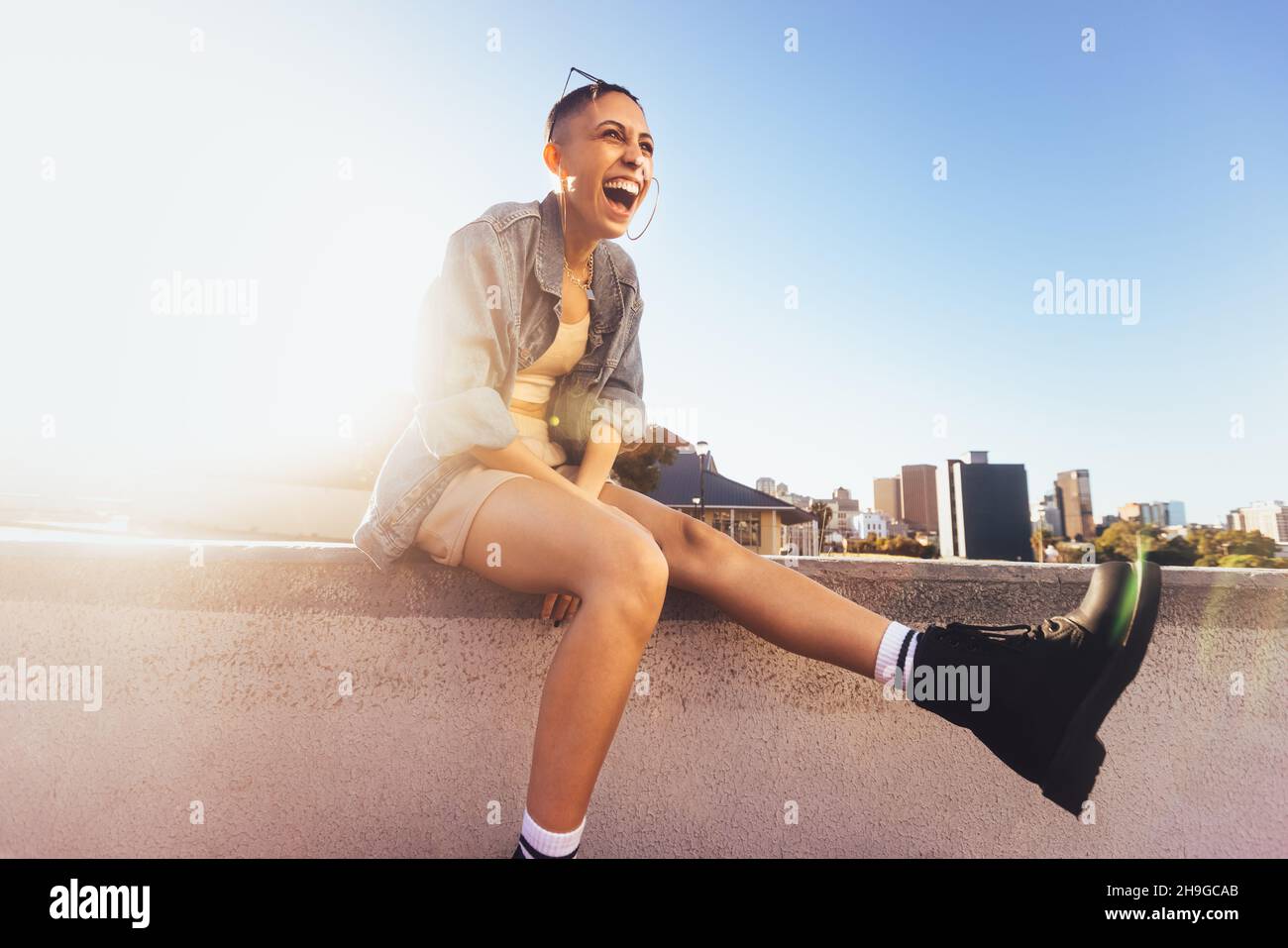 Hochgelaunte junge Frau lacht fröhlich, während sie an einer Wand in der Sonne sitzt. Glückliche junge Frau, die sich lebhaft und voller Leben in der Stadt fühlt. Stockfoto