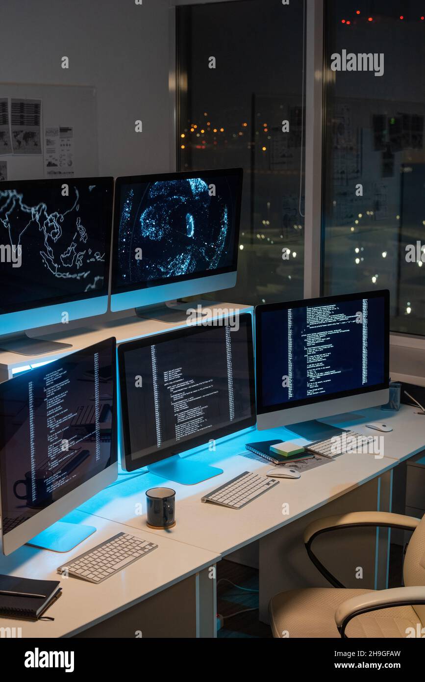 Gruppe von Computermonitoren mit codierten Daten, die nachts auf Schreibtischen im leeren Büro des Programmierunternehmens stehen Stockfoto