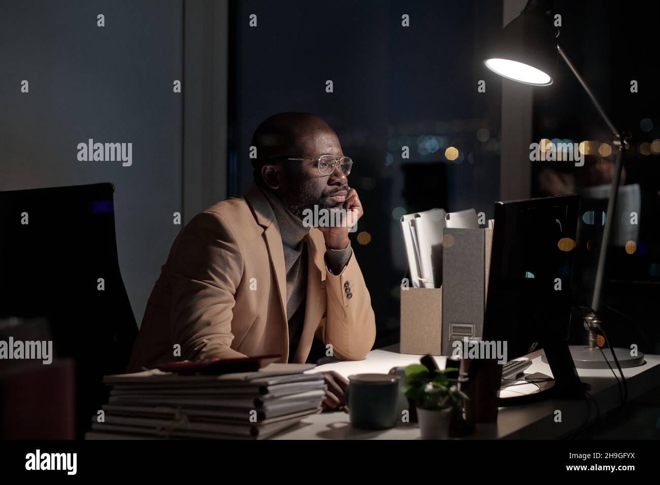 Afrikanischer Geschäftsmann, der am Schreibtisch vor einem Computermonitor im Büro brummend arbeitet, während er spät in der Nacht arbeitet Stockfoto