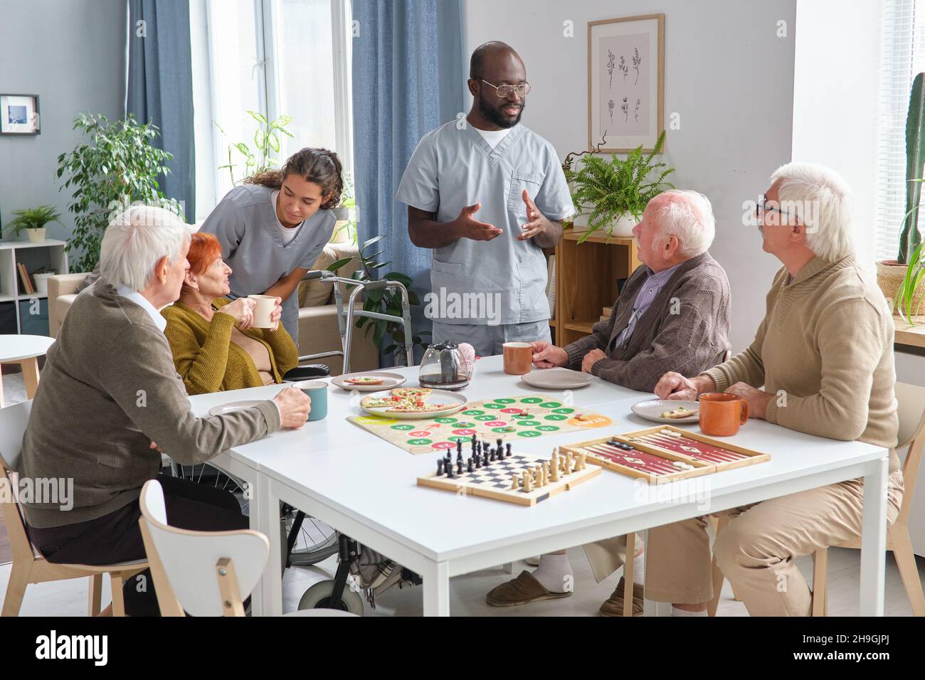 Gruppe von Senioren spielen Brettspiel am Tisch und sprechen mit medizinischem Personal im Zimmer des Pflegeheims Stockfoto