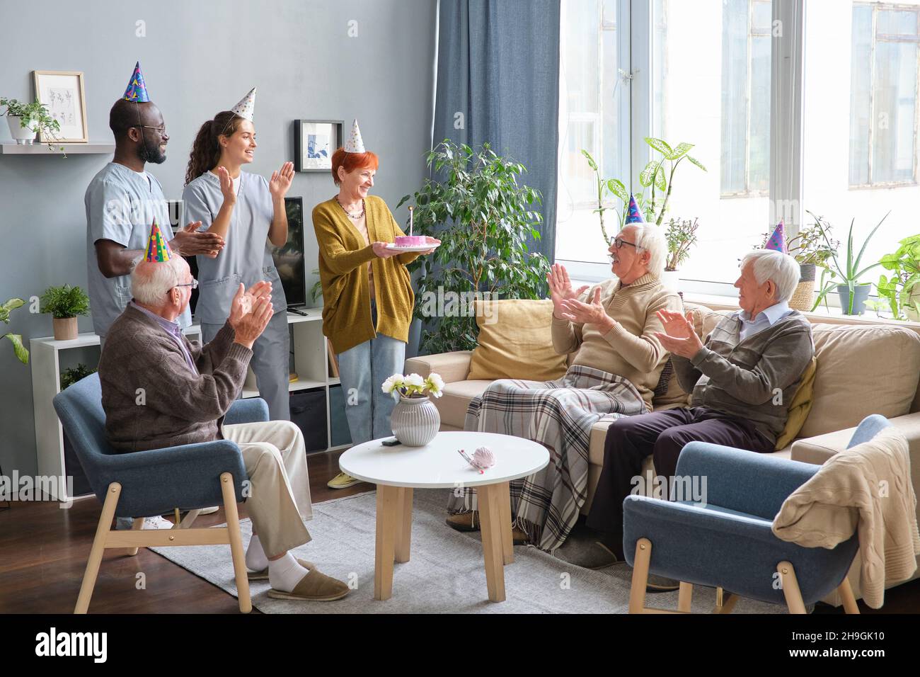 Ältere Menschen mit medizinischem Personal klatschen sich die Hände und gratulieren der Frau zum Geburtstag, während sie den Geburtstagskuchen im Zimmer hielt Stockfoto