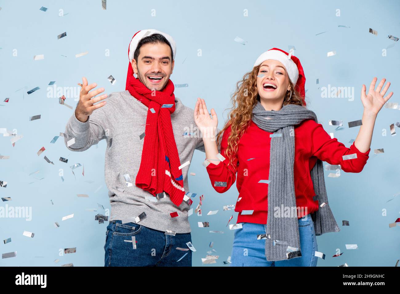 Glückliches kaukasisches Paar lächelt und hebt die Hände hoch und feiert Weihnachten im hellblauen Studiohintergrund mit Konfetti Stockfoto