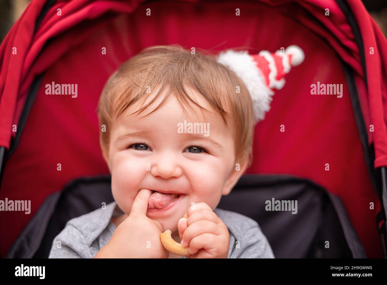 Baby sitzt im roten Kinderwagen. Das Kind isst einen Bagel und zeigt seine Zunge. Stockfoto