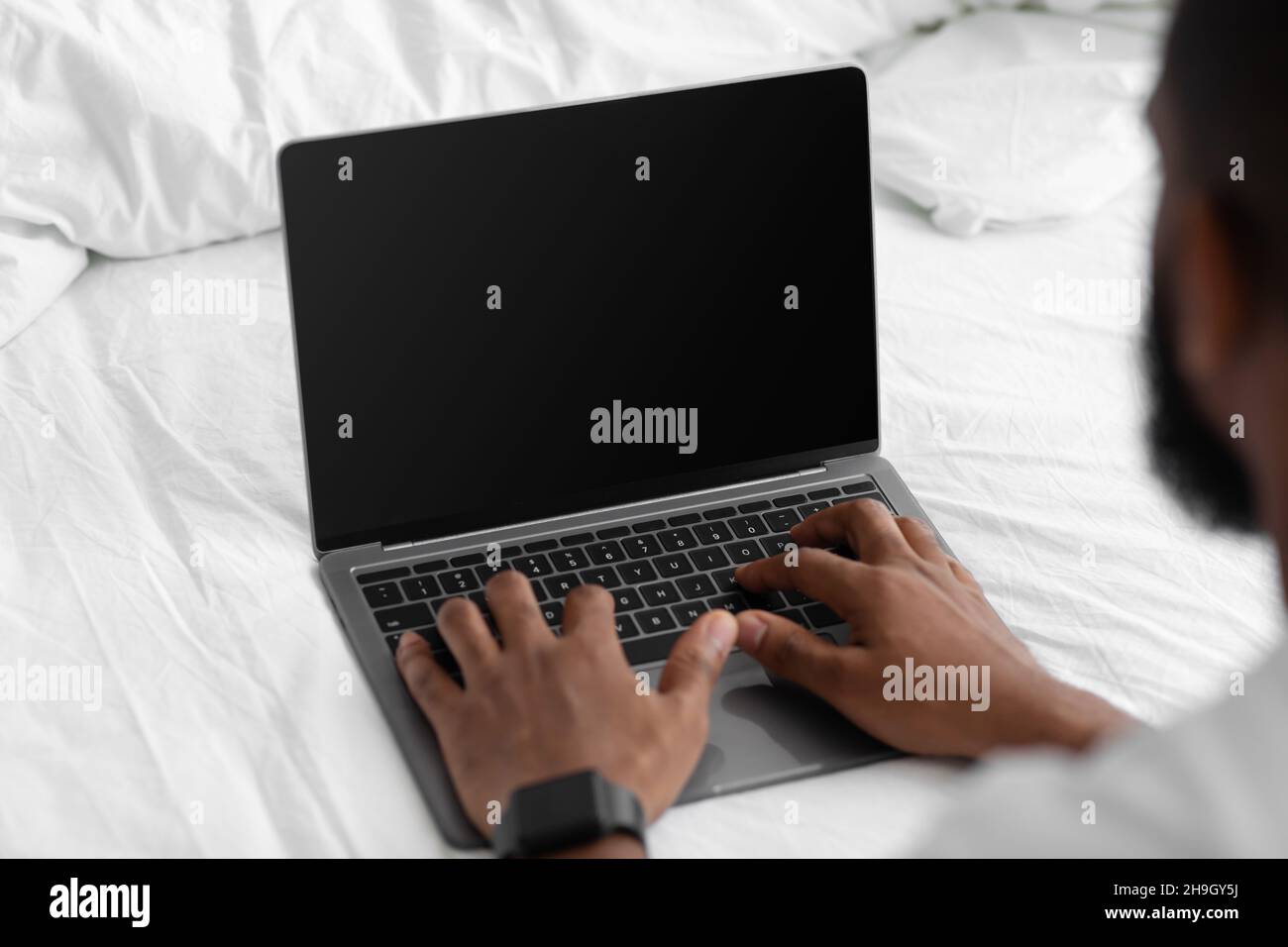 Der junge afroamerikanische Mann schaut sich den Laptop mit leerem Bildschirm an, arbeitet im Schlafzimmer fern auf dem weißen Bett Stockfoto