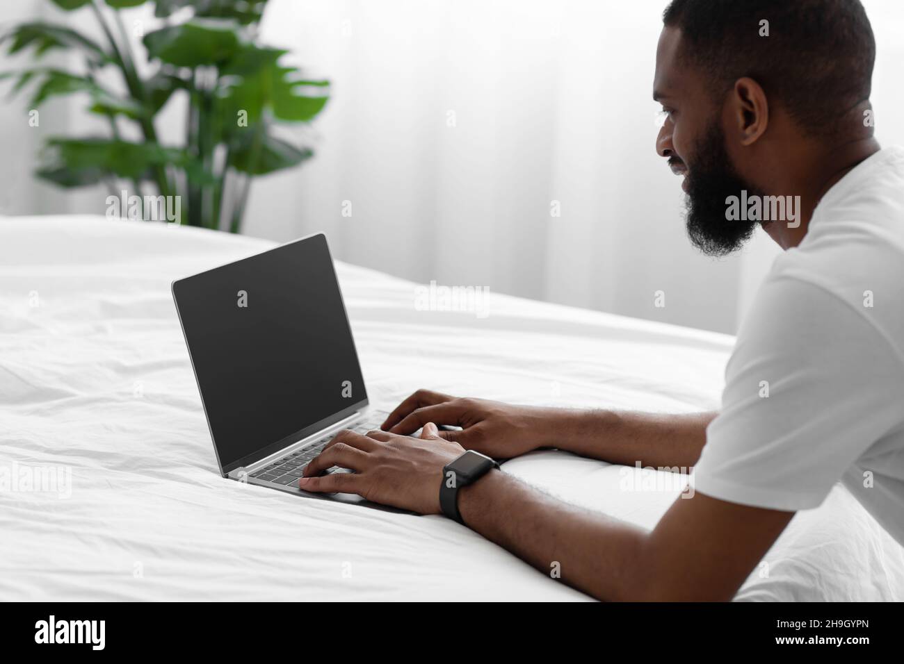 Lächelnder, tausendjähriger afroamerikanischer bärtiger Mann blickt auf einen Laptop mit leerem Bildschirm und arbeitet fernab auf einem weißen Bett Stockfoto