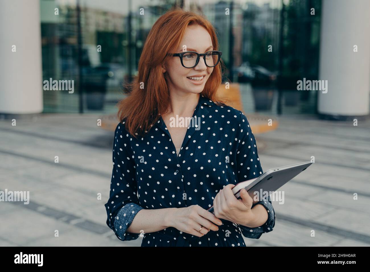 Das weibliche Modell trägt einen Laptop und der Organisator posiert draußen und wartet auf jemanden in der Nähe des Bürogebäudes Stockfoto