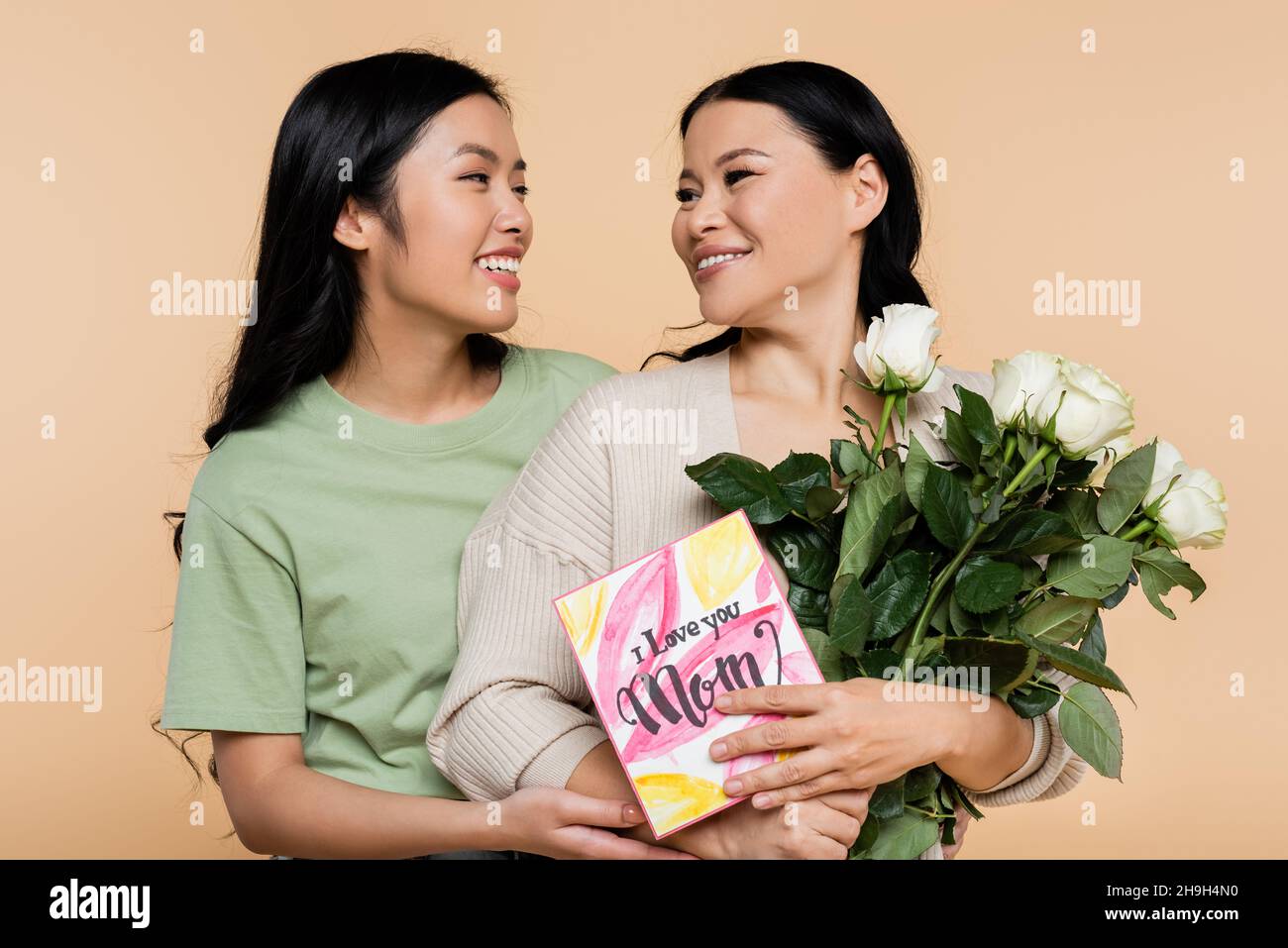 Fröhliche asiatische Frau umarmt Mutter mit Rosen und Grußkarte isoliert auf Beige Stockfoto