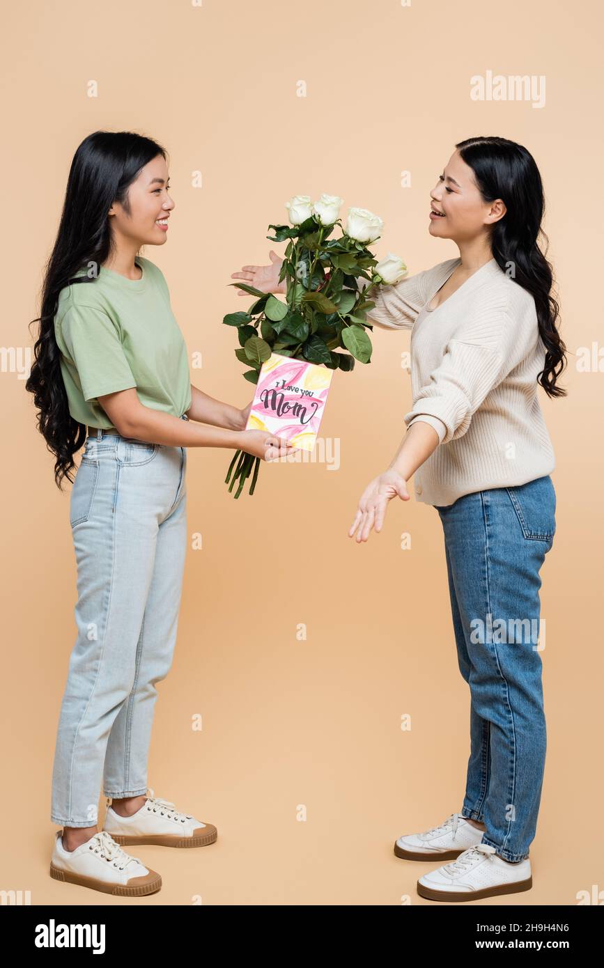 Glückliche asiatische Tochter gratuliert Mutter mit Grußkarte und Blumen auf beige Stockfoto