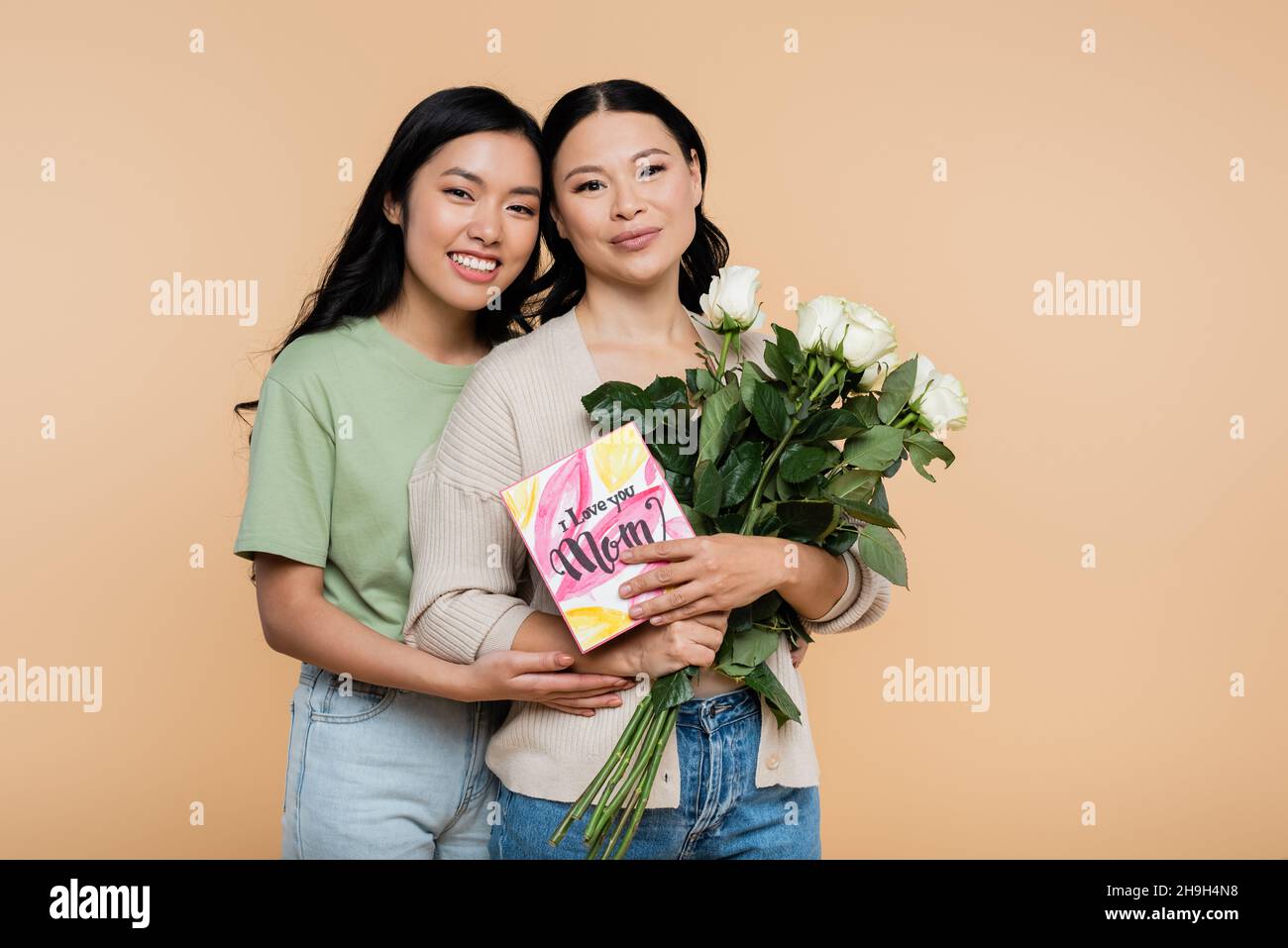 Glückliche asiatische Frau umarmt Mutter mit Blumen und Grußkarte isoliert auf beige Stockfoto