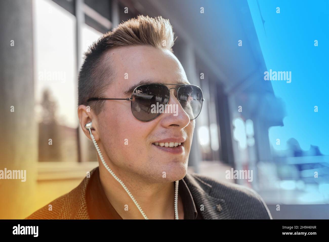 Porträt eines positiv lächelnden jungen Geschäftsmannes mit europäischem Auftritt in Sonnenbrillen und Kopfhörern, der draußen auf der Straße Musik hört. Stockfoto